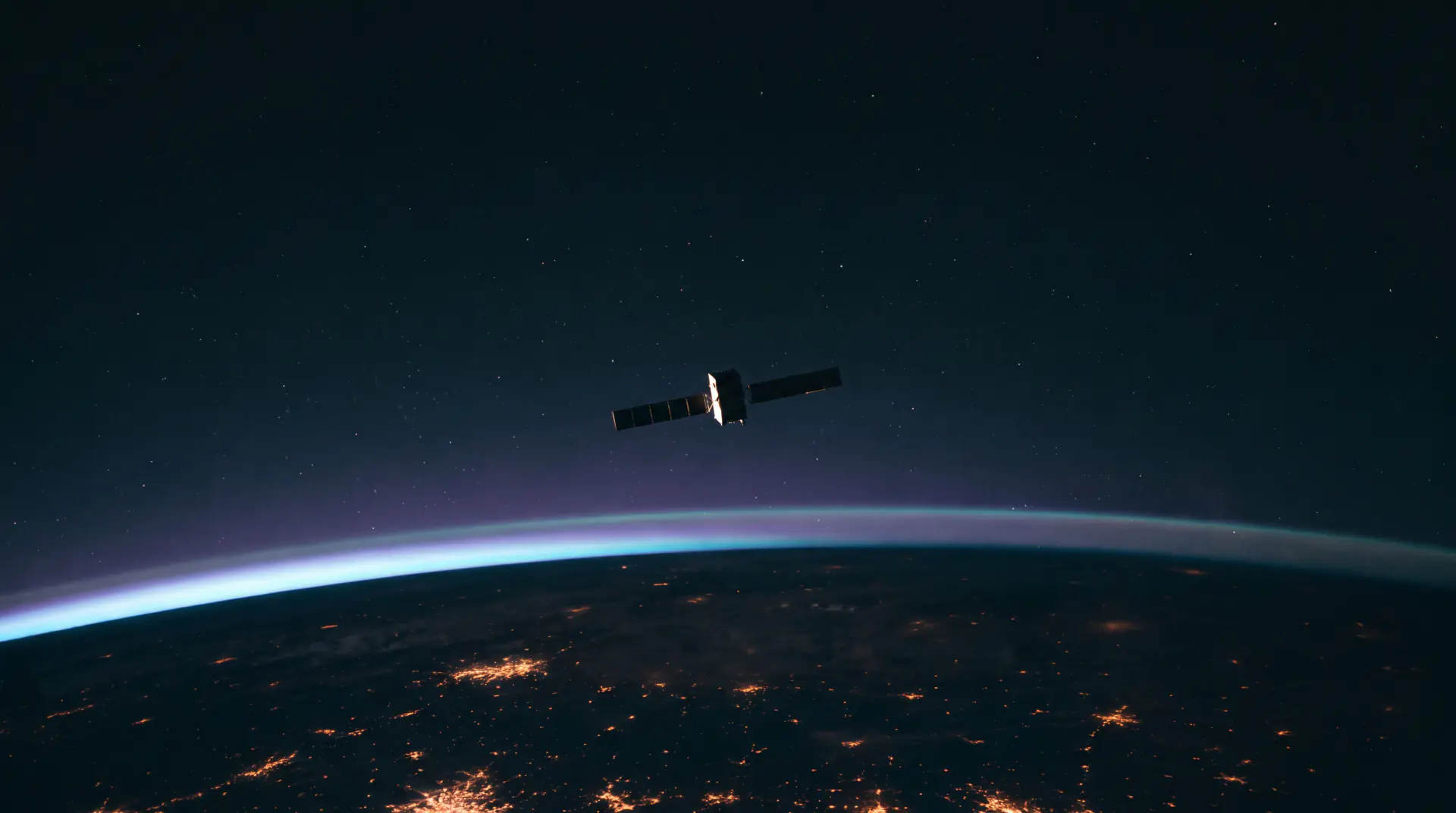A satellite in silhouette above the night side of Earth, the atmosphere a thin lit band below it.